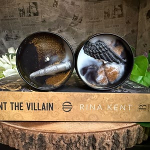 May include: Two black candles with decorative elements, one with a silver object and gold glitter, the other with a black wing and gold glitter. The candles sit on a book titled "Hunt the Villain" by Rina Kent.