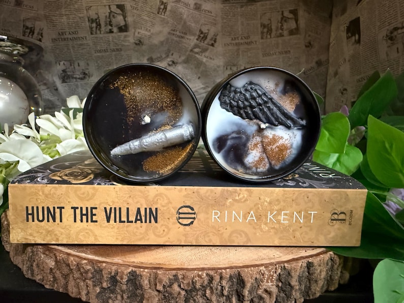 May include: Two black candles with decorative elements, one with a silver object and gold glitter, the other with a black wing and gold glitter. The candles sit on a book titled "Hunt the Villain" by Rina Kent.