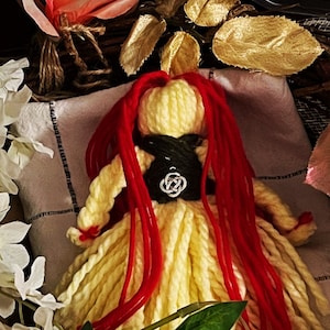 May include: A yellow yarn doll with long red yarn hair. The doll is wearing a black and green vest with a silver Celtic knot pendant. The doll is surrounded by white and pink flowers.
