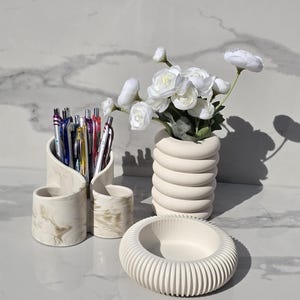 May include: A set of cream-coloured desk accessories. Includes a pen holder with pens, a ribbed bowl, and a vase with white flowers. The pen holder has a marble design. The vase has a stacked, cylindrical design.