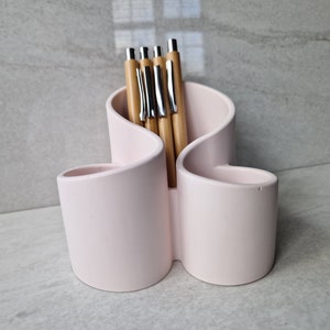 May include: A pale pink desk tidy with three sections, containing several wooden pens with silver detailing. The organiser has a distinctive, undulating design and is set against a light grey backdrop.