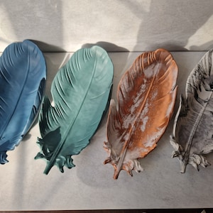 May include: Four decorative ceramic feather trays in blue, teal, brown, and grey. The trays are shaped like feathers and have a textured finish.