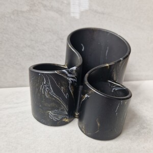 May include: A black and gold marbled decorative container with three compartments. The container has a unique, wavy design and is made of a solid material. The interior of the container is black, and the exterior has gold and white marbling.