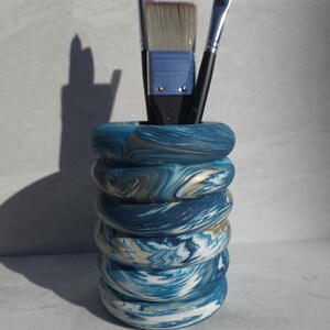 May include: A decorative art supply holder with a swirling blue, white, and gold pattern. The holder contains several paint brushes with black handles and light-coloured bristles. The holder is cylindrical and stacked with ring-shaped layers.