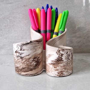May include: A white ceramic pen holder with a brown marbled design. The holder has two compartments and is filled with colourful pens, including pink, blue, green, and yellow.