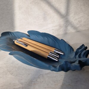 May include: Four wooden pens with silver accents are arranged in a blue feather-shaped dish.