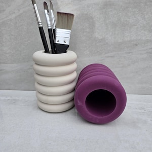 May include: Two cylindrical brush holders, one white and one purple. The white holder has a stacked ring design and holds several paintbrushes. The purple holder is solid and has a smooth surface.