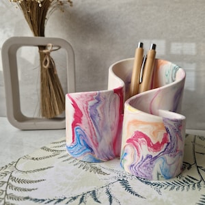 May include: A colourful ceramic desk organiser with a marbled design in shades of pink, blue, and yellow. The organiser has three compartments and is holding two wooden pens.