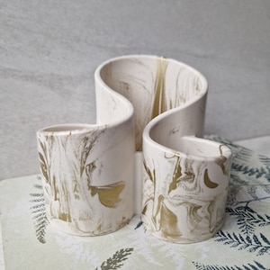 May include: White ceramic holder with a marbled design and gold accents. The holder has three sections, each with a curved shape.