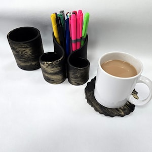 May include: A white coffee mug with a brown liquid sits on a black and gold coaster. A black pen holder with gold accents holds colourful pens. A matching black container is to the left.