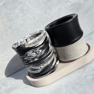 May include: Two ceramic planters on a beige tray. One has a black and white marbled design, the other is black on top with a dotted pattern on the bottom. The tray is oval-shaped.