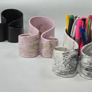 May include: Three decorative desk organisers in different colours and patterns. The organisers are made of a smooth, ceramic-like material and have a unique, wavy design. The organisers are perfect for holding pens, pencils, and other office supplies.