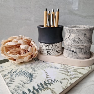 May include: Two ceramic pencil holders, one black with white polka dots and one white with grey marbling. The holders are on a tray with a fern pattern and a light pink flower.