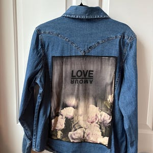 May include: A blue denim jacket with a rectangular graphic on the back. The graphic features the word "LOVE" in white text above a floral design with pink and white flowers. The word "AMOUR" is printed in reverse.