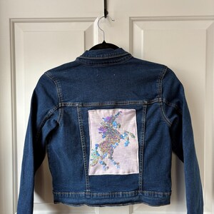May include: A dark blue denim jacket with a light pink patch on the back. The patch features a sequined unicorn design in shades of purple, blue, and silver. The jacket is hanging on a black hanger.
