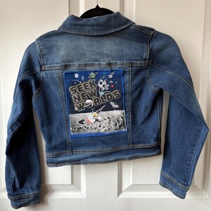 May include: Blue denim jacket with a patch on the back that says "Seek New Worlds." The patch features a rocket ship, planets, and a lunar landscape. The jacket has a classic collar and button closures.