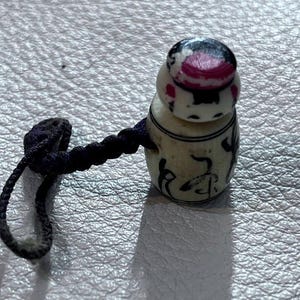 May include: A small, off-white decorative container with a dark purple cord. The container has a rounded top with a black and pink design. The body of the container has black painted details.