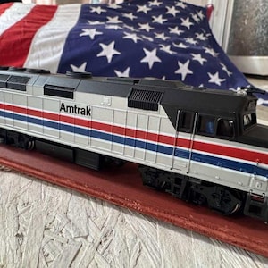 Puede incluir: Una locomotora de tren Amtrak modelo con rayas plateadas, rojas y azules. El número 217 es visible en el lateral. El tren está sobre una superficie roja, con una bandera estadounidense de fondo.