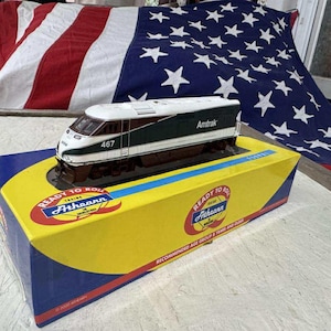 May include: A model Amtrak train, white and green with the number 467, sits atop a yellow and blue box. The box has the words "Ready to Roll" and "Athearn" printed on it. An American flag is in the background.