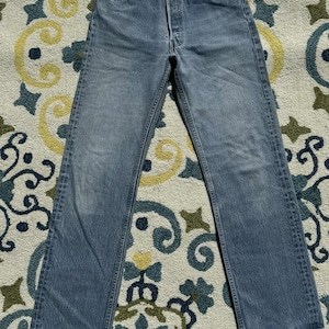 Puede incluir: Vaqueros de mezclilla azul claro con bragueta de botones. Los vaqueros tienen un diseño clásico de cinco bolsillos y un corte recto. La mezclilla tiene un aspecto descolorido, lo que sugiere desgaste y lavado. Los vaqueros están colocados planos sobre una superficie estampada.