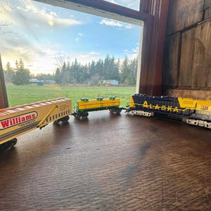 May include: A model train set with a yellow and black Alaska locomotive, a yellow tank car, and a yellow Williams boxcar. The train cars are on a wooden surface, with a window and a landscape in the background.