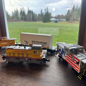 Op de afbeelding: Een verzameling modeltreinwagons, waaronder een gele Union Pacific locomotief en caboose, een platte wagen met een witte container en een caboose met een patriottisch ontwerp. De modellen worden op een houten oppervlak gepresenteerd.