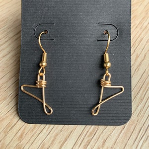 May include: A pair of gold-colored wire earrings shaped like miniature clothes hangers. The earrings are displayed on a black earring card with a small pink circle at the top. The background is a light wood grain.
