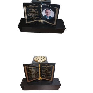May include: A black memorial stone shaped like an open book, with gold lettering and a photograph. The text reads "Cherished Memories" and includes a photo of a man. A gold-coloured flower holder is on top.