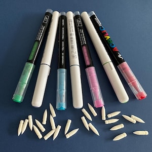 May include: Six white acrylic markers with different coloured tips, including green, blue, turquoise, pink, and red. The markers are arranged in a fan shape with the tips pointing towards the bottom of the image. There are also 20 white paint brush tips scattered around the markers.
