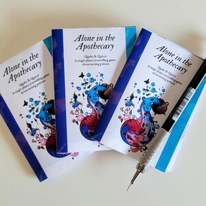 Może przedstawiać: Trzy egzemplarze gry "Alone in the Apothecary" z niebiesko-czerwono-białą okładką. Gra polega na mieszaniu mikstur. Czarny i srebrny oł&oacute;wek mechaniczny spoczywa na książkach.
