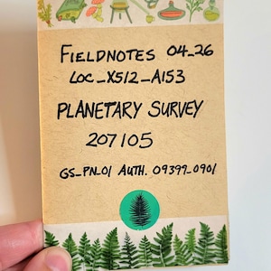 May include: A small, rectangular notebook with the text "FIELDNOTES 04-26", "PLANETARY SURVEY", and other details. The notebook has a decorative border with illustrations of plants and tools. A green sticker with a fern is visible.