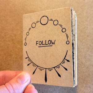 May include: A small, brown paper notebook with a black circle design and the word "FOLLOW" written in the center. The notebook is closed and has a hand holding it.