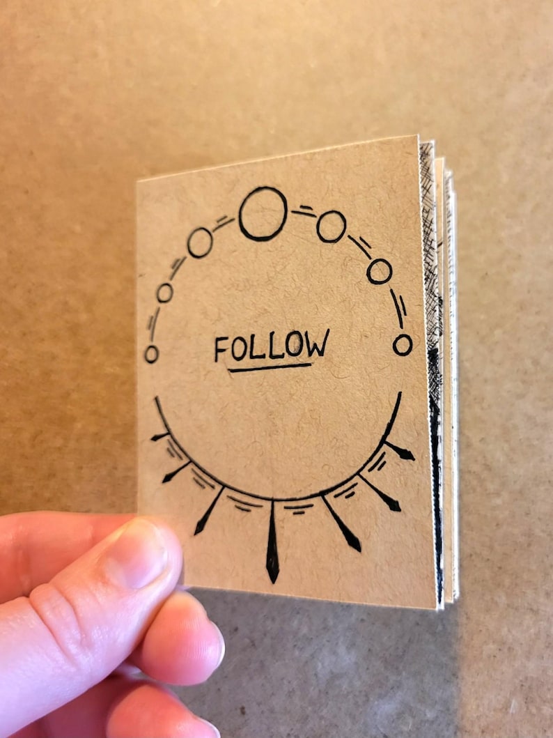 May include: A small, brown paper notebook with a black circle design and the word "FOLLOW" written in the center. The notebook is closed and has a hand holding it.