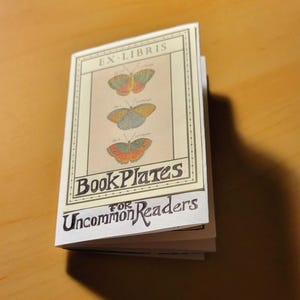 May include: A small book with a white cover and a black border. The cover features a vintage illustration of three butterflies and the text "Ex-Libris Book Plates for Uncommon Readers".