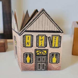 Pode incluir: Uma casa de papel com telhado marrom e janelas amarelas. As janelas t&ecirc;m placas que dizem "IN THIS HOUSE".