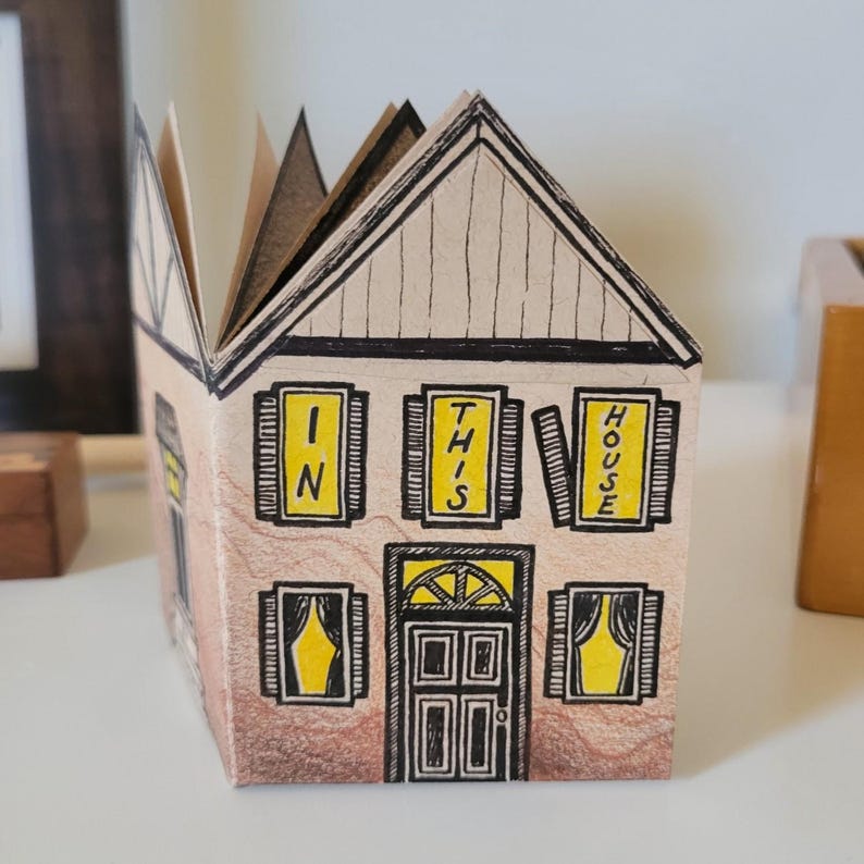 Pode incluir: Uma casa de papel com telhado marrom e janelas amarelas. As janelas t&ecirc;m placas que dizem "IN THIS HOUSE".
