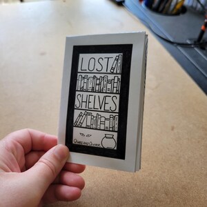 May include: A black and white illustration of a bookshelf with the words "LOST SHELVES" above it. The illustration includes a pair of eyes peeking out from below the bottom shelf and a jar with a lid on top of the shelf. The text "Quirk and Quimer" is below the illustration.