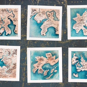 May include: Six watercolor map prints, each framed in white. The maps depict landmasses with mountains, rivers, and coastlines, rendered in shades of brown and blue. The background is a dark, patterned surface.