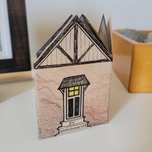 Pode incluir: Uma pequena casa de papel com uma ilustra&ccedil;&atilde;o em preto e branco de uma janela e um telhado. A casa &eacute; decorada com linhas marrons e pretas e o texto "BY QUAKEANDQUIVER".