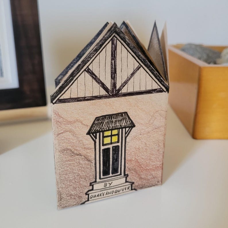 Pode incluir: Uma pequena casa de papel com uma ilustra&ccedil;&atilde;o em preto e branco de uma janela e um telhado. A casa &eacute; decorada com linhas marrons e pretas e o texto "BY QUAKEANDQUIVER".