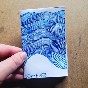 Puede incluir: Un pequeño folleto de papel plegado con un diseño de olas azules. Las olas están creadas con líneas finas y paralelas. El texto "SEA-FEVER" está impreso en azul en la parte inferior. El folleto está sostenido por una mano.