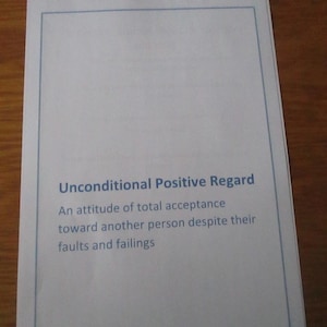 May include: A white rectangular paper with the text "Unconditional Positive Regard" and a definition in blue font. The paper is on a wooden surface.