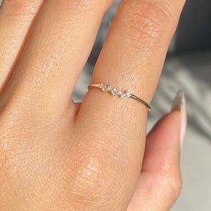 May include: A delicate gold ring featuring five small, clear gemstones arranged in a row. The ring has a thin, minimalist design, suitable for wearing on a finger. The gemstones sparkle in the light.