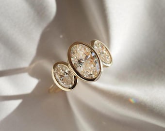 Può includere: Un anello d'oro con tre diamanti taglio ovale. I diamanti sono incastonati in una montatura a castone e l'anello è esposto su uno sfondo bianco e setoso. Il design dell'anello è elegante e moderno, mettendo in risalto la brillantezza.