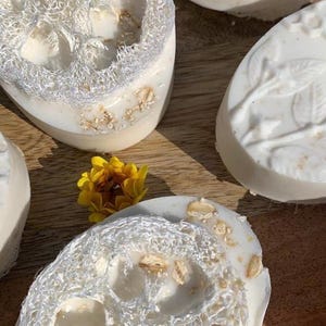 K&ouml;nnte beinhalten: Nahaufnahme von handgemachten Seifen mit Luffa und floralen Akzenten. Die wei&szlig;en Seifen enthalten Luffa und Haferflocken. Eine gelbe und eine rote Blume liegen auf einer Holzoberfl&auml;che. Die Seifen haben eine nat&uuml;rliche, spa-&auml;hnliche &Auml;sthetik.