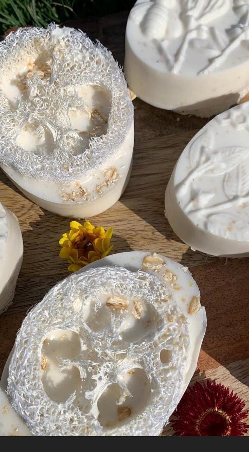 K&ouml;nnte beinhalten: Nahaufnahme von handgemachten Seifen mit Luffa und floralen Akzenten. Die wei&szlig;en Seifen enthalten Luffa und Haferflocken. Eine gelbe und eine rote Blume liegen auf einer Holzoberfl&auml;che. Die Seifen haben eine nat&uuml;rliche, spa-&auml;hnliche &Auml;sthetik.