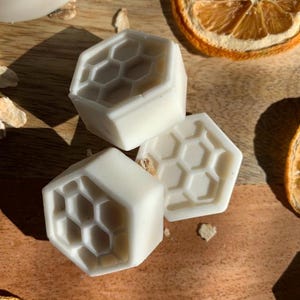 K&ouml;nnte beinhalten: Drei wei&szlig;e, sechseckige Seifen mit einem Wabenmuster liegen auf einer Holzoberfl&auml;che, zusammen mit getrockneten Orangenscheiben. Die Seifen haben eine saubere, minimalistische &Auml;sthetik, die auf ein nat&uuml;rliches Hautpflegeprodukt hindeutet.