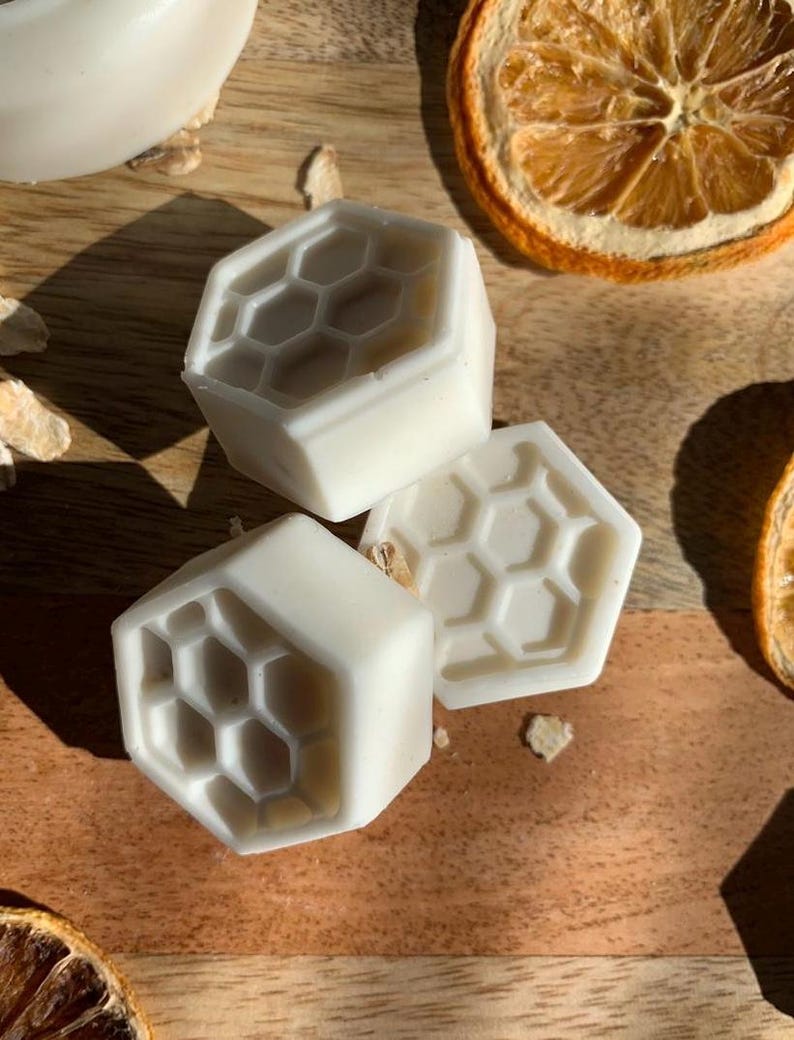 K&ouml;nnte beinhalten: Drei wei&szlig;e, sechseckige Seifen mit einem Wabenmuster liegen auf einer Holzoberfl&auml;che, zusammen mit getrockneten Orangenscheiben. Die Seifen haben eine saubere, minimalistische &Auml;sthetik, die auf ein nat&uuml;rliches Hautpflegeprodukt hindeutet.