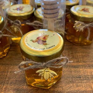 May include: Small glass jars filled with honey, tied with twine and a gold bee charm. Each jar has a Winnie the Pooh themed label that reads "A little yummy for your tummy".