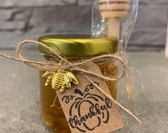 Könnte beinhalten: Ein kleines Glas mit Honig, mit einem goldenen Deckel und einem dekorativen Bienenanhänger. Ein Etikett mit dem Wort "thankful" ist mit Bindfaden befestigt. Ein hölzerner Honiglöffel ist enthalten.